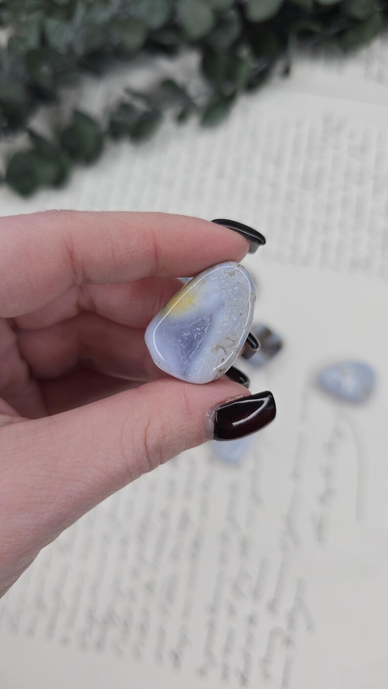 Puede incluir: Una piedra preciosa de &aacute;gata pulida, de color azul, con inclusiones amarillas y blancas. La piedra se sostiene en una mano, con otras piedras similares visibles en el fondo. El &aacute;gata tiene una forma suave y redondeada.