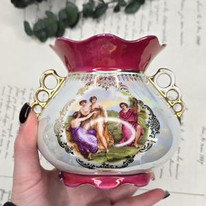 Peut inclure: Un vase en porcelaine avec un bord et une base festonnés magenta, orné d'une décalcomanie centrale représentant trois personnages en tenue classique dans un cadre en filigrane doré. Le corps est blanc irisé avec deux anses dorées ajourées.