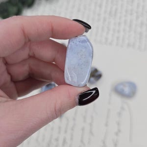Puede incluir: Una piedra rodada azul claro con inclusiones blancas y grises. La piedra se sostiene entre los dedos, con otras piedras visibles en el fondo. La piedra es lisa y pulida, con una forma ligeramente redondeada. El fondo es claro.