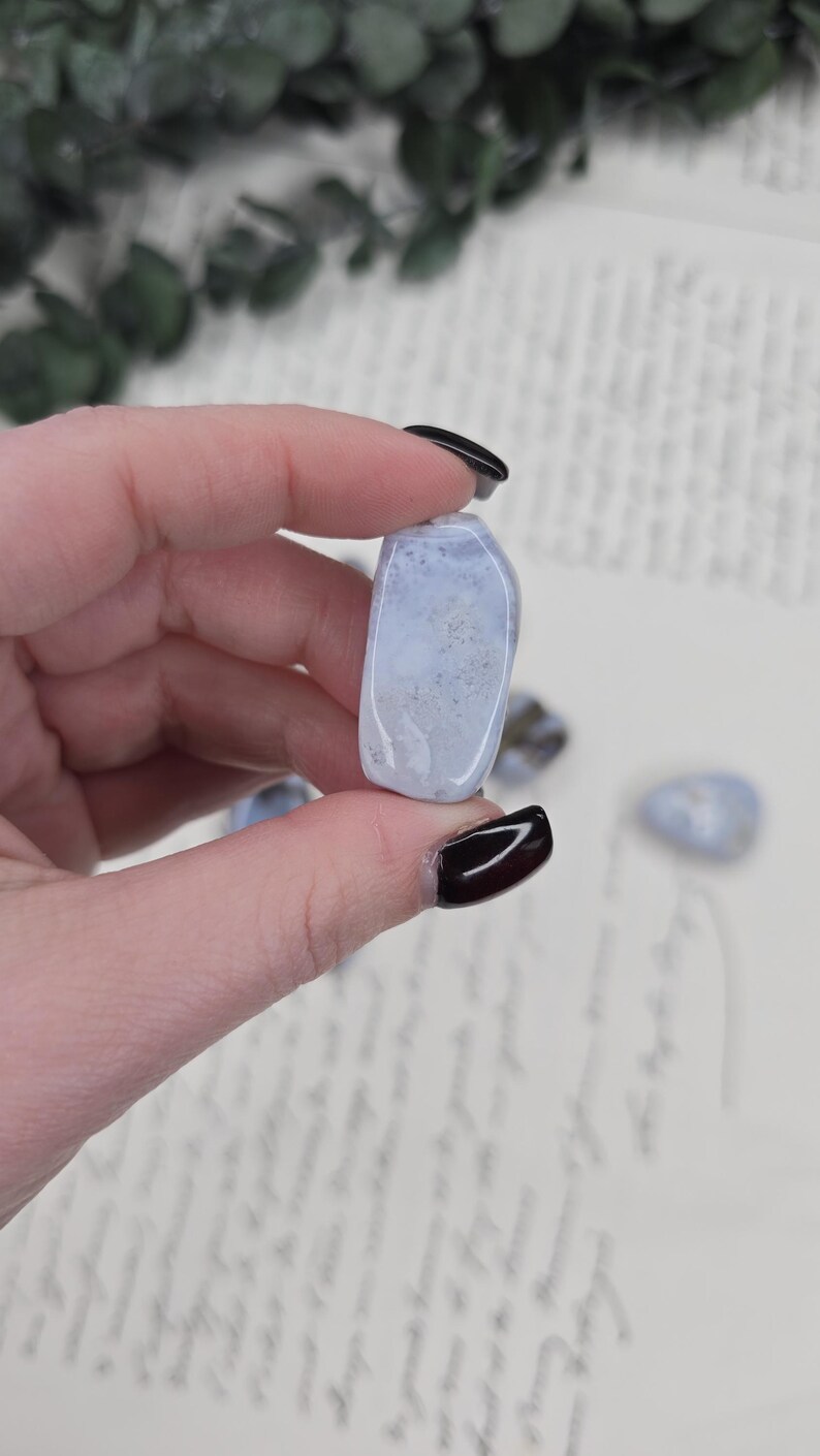 Puede incluir: Una piedra rodada azul claro con inclusiones blancas y grises. La piedra se sostiene entre los dedos, con otras piedras visibles en el fondo. La piedra es lisa y pulida, con una forma ligeramente redondeada. El fondo es claro.