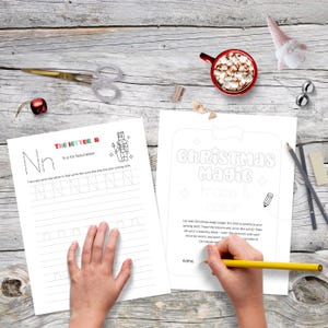 May include: A festive scene with two activity sheets featuring the text "The Letter N" and "Christmas Magic". A hand holds a yellow pencil, coloring the "Christmas Magic" sheet. Other items include scissors, a mug of cocoa, and holiday decorations.