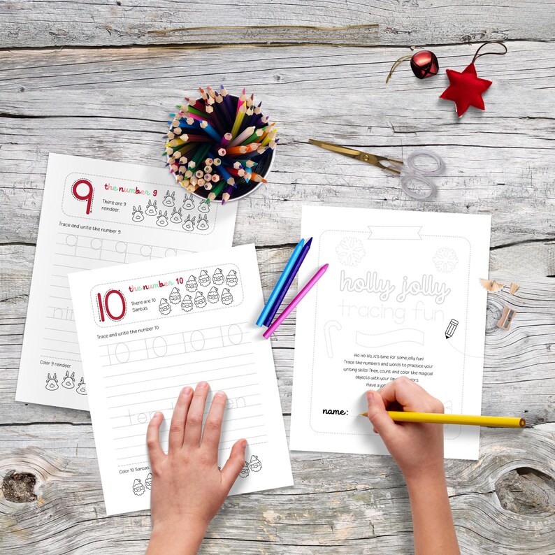May include: A collection of Christmas-themed tracing and coloring pages with the numbers 9 and 10. A hand is holding a yellow pencil, tracing the words "Holly Jolly tracing fun". Colored pencils and scissors are also on the wooden surface.