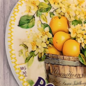 May include: Round decorative ceramic coaster featuring a vibrant illustration of lemons and yellow flowers spilling from a wooden barrel. The word "PORTUGAL" is prominently displayed in purple. The coaster has a yellow stitched border.
