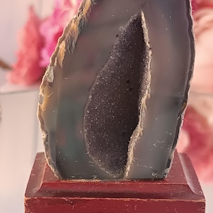 Peut inclure: Une tranche de géode d'agate gris foncé polie, avec un intérieur cristallin, montée sur un petit socle rectangulaire en bois brun rougeâtre. Le bord extérieur de la géode est de couleur beige clair. L'arrière-plan est flou avec des tons roses et blancs.