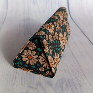 Pode incluir: Um estojo triangular para óculos com fundo preto e um padrão floral de margaridas em tons de bege, laranja e verde-azulado. O estojo está fechado e parece ser feito de um material texturizado.