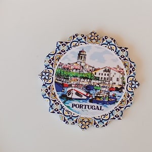 Pode incluir: Um ímã redondo e colorido com uma imagem de uma cidade e um porto português. O ímã tem uma borda com padrão azul, branco e amarelo e a palavra "PORTUGAL" impressa em preto na parte inferior.