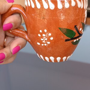 Pode incluir: Caneca de terracota artesanal com alça, decorada com pontos brancos e detalhes de folhas verdes. A borda apresenta um design branco ondulado. A caneca é segurada por uma mão com esmalte rosa. Uma peça única.
