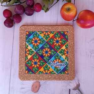 Könnte beinhalten: Quadratischer Korkuntersetzer mit einem farbenfrohen Blumenmuster in Rot, Gelb, Blau und Grün. Das Wort "Portugal" ist auf dem Untersetzer geschrieben. Umgeben von Pflaumen, Äpfeln und Grün auf einer weißen Holzoberfläche.