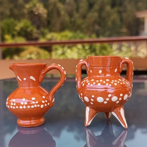 Puede incluir: Dos pequeñas piezas de cerámica marrón con lunares blancos. Una es una jarra con asa y la otra es una olla con tres patas.