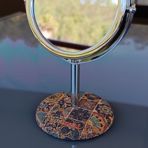 Pode incluir: Um espelho de penteadeira com moldura prateada e superfície redonda e reflexiva. O espelho está em um suporte com uma base colorida e estampada. A base tem a palavra "Portugal" impressa.