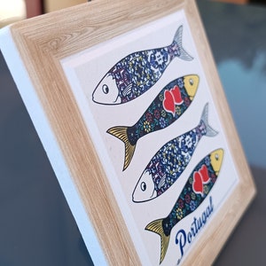 Pode incluir: Um azulejo emoldurado com fundo branco e moldura de madeira em tom de madeira. O azulejo apresenta três peixes coloridos com padrões azuis, amarelos, vermelhos e brancos. A palavra "Portugal" está escrita em itálico na parte inferior do azulejo.