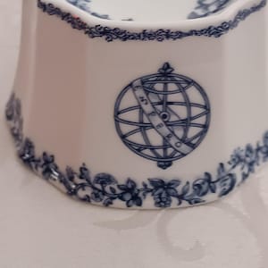 Può includere: Posacenere in ceramica bianca con disegni floreali e globo blu. Il posacenere ha una forma svasata con bordo decorativo ed emblema del globo. Il bordo e la base presentano motivi floreali blu.
