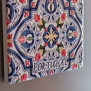 Può includere: Una piastrella di ceramica bianca con un motivo floreale blu, rosso, giallo e verde. La piastrella ha la scritta "PORTUGAL" in blu nella parte inferiore.