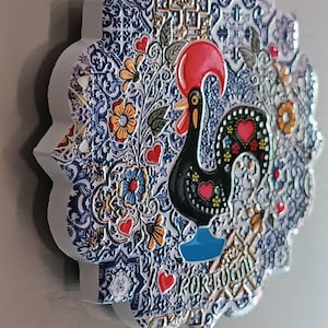 Pode incluir: Um ímã de cerâmica branca com um design de padrão azul e branco que apresenta um galo preto com acentos vermelhos. O galo está em pé sobre uma base azul e a palavra "PORTUGAL" está escrita em letras douradas abaixo do galo.