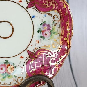 Puede incluir: Primer plano de un platillo de porcelana con diseño floral. El platillo tiene una base blanca con detalles rosas y dorados. Flores pintadas a mano y filigranas doradas adornan el borde. El platillo está sostenido por un soporte de metal color bronce.
