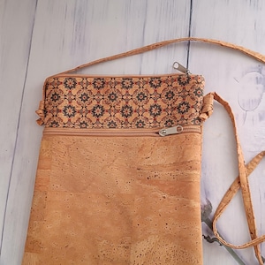 Pode incluir: Uma bolsa tiracolo retangular feita de material de cortiça castanho claro. A secção superior apresenta uma faixa decorativa com um padrão floral repetitivo em azul escuro e vermelho. Inclui uma alça de cortiça fina e um fecho de correr prateado.