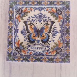 Pode incluir: Azulejo cerâmico quadrado com um desenho de borboleta. A borboleta tem asas azuis, laranja e pretas. O azulejo tem uma borda floral azul e laranja e a palavra "PORTUGAL" está escrita no azulejo.
