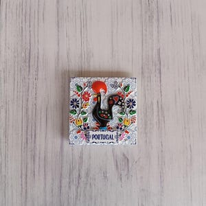 Pode incluir: Um azulejo de cerâmica branco com um desenho de galo colorido. O galo é preto com crista e barbelas vermelhas. O azulejo é rodeado por uma borda floral e a palavra "PORTUGAL" está impressa no centro inferior.