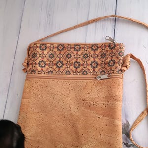 Pode incluir: Uma bolsa tiracolo retangular feita de material de cortiça em tom castanho natural. A secção superior apresenta um padrão decorativo de pequenos motivos florais em azul escuro e vermelho. Inclui um bolso horizontal com fecho e uma alça fina.