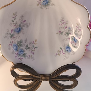 Pode incluir: Um prato de porcelana branca em forma de concha com delicados padrões florais de rosas azuis, folhas verdes e detalhes roxos. O prato tem uma borda recortada com acabamento dourado e está exposto num suporte de metal cor de bronze em forma de laço.