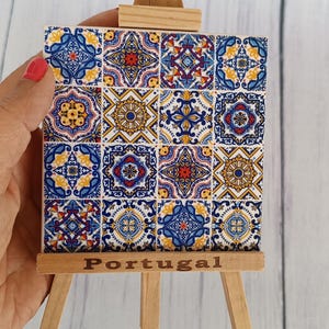 Pode incluir: Um pequeno cavalete de madeira exibindo um azulejo com um design tradicional português. O azulejo apresenta um padrão repetitivo de motivos florais azuis, amarelos e vermelhos. A palavra "Portugal" está escrita na base do cavalete.