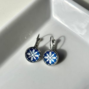 Könnte beinhalten: Ein Paar silberne Ohrhänger mit blauen und weißen gemusterten Glascabochons. Das Muster ist ein weißes Schneeflocken-Design auf einem dunkelblauen Hintergrund.