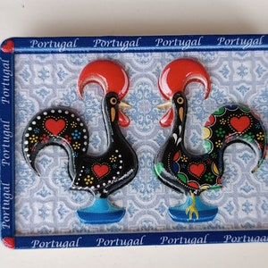 May include: A blue and white ceramic tile magnet featuring two black roosters with red combs and colourful designs. The roosters are facing each other and the tile is surrounded by a blue border with the word "Portugal" repeated around the edge.
