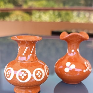 Pode incluir: Dois pequenos vasos de terracota com desenhos pintados de branco. O vaso à esquerda tem um padrão circular com pontos brancos, enquanto o vaso à direita tem um desenho mais abstrato com pontos e espirais brancos.