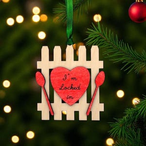 May include: Wooden Christmas ornament featuring a white picket fence design with a red heart in the center. The heart has the text "I'm Locked in". Two red spoons flank the heart. A green ribbon is attached for hanging.