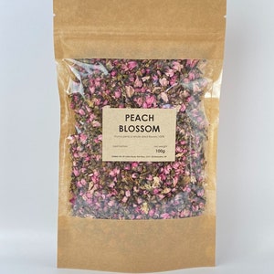 May include: A brown paper bag with a clear plastic window, containing pink and green dried peach blossoms. The bag has a label that reads "Peach Blossom" and "Prunus persica whole dried flowers 100%". The label also includes "Net weight 100g".