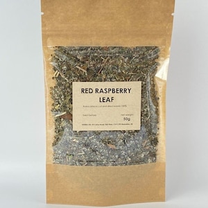 May include: A brown paper bag with a clear window, filled with dried red raspberry leaf. A white label on the bag reads "RED RASPBERRY LEAF" and "net weight 50g". The bag is sealed with a zip closure.