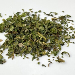 May include: A pile of dried, green, leafy herbs. The leaves vary in size and shape, with some appearing whole and others fragmented. The color ranges from vibrant green to a muted, brownish-green, suggesting varying levels of dryness. The herbs are scattered on a white surface.