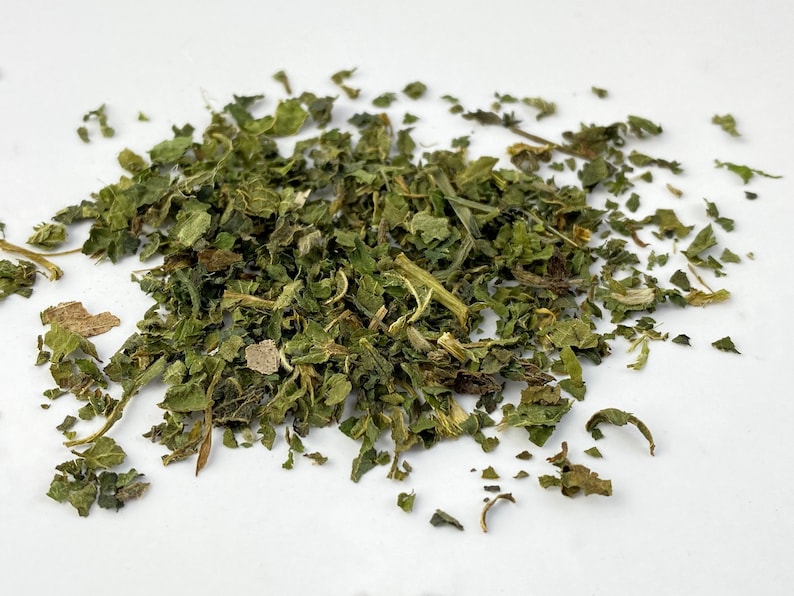 May include: A pile of dried, green, leafy herbs. The leaves vary in size and shape, with some appearing whole and others fragmented. The color ranges from vibrant green to a muted, brownish-green, suggesting varying levels of dryness. The herbs are scattered on a white surface.