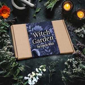 May include: A brown cardboard box with a blue floral label reading "Witch Garden The Eclectic Witch." The box is surrounded by herbs, flowers, candles, and a mortar and pestle on a dark surface.