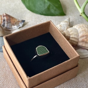 May include: A green sea glass ring in a brown cardboard box with a black interior. The ring is set in silver.