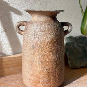 Puede incluir: Un jarrón de cerámica texturizada hecho a mano en tonos marrones y beige. El jarrón tiene un cuerpo cilíndrico con dos pequeñas asas de bucle y un borde acampanado. La cerámica rústica es adecuada para la decoración del hogar.