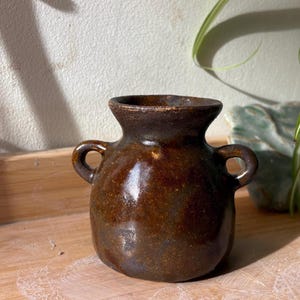 May include: A small, handmade brown ceramic vase with two loop handles. The vase has a rounded body and a slightly flared rim. The glaze has a glossy finish. The vase is a decorative item, likely for displaying flowers or other small items.