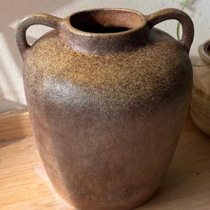 Peut inclure: Un grand vase en céramique marron avec deux anses. Le vase a un corps large et arrondi et une petite ouverture en haut. Le dégradé de couleurs passe d'un beige clair à un marron plus foncé.