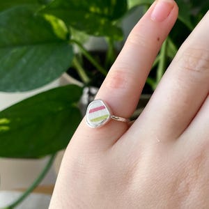Könnte beinhalten: Ein silberner Ring mit einem weißen Stein, der mit drei rosa und grünen Streifen verziert ist.
