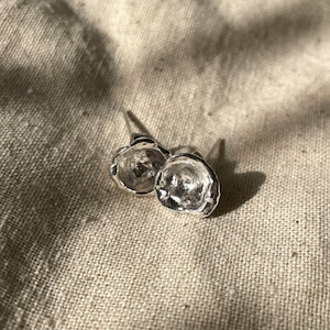 Puede incluir: Un par de pendientes de plata con una forma orgánica texturizada. Los pendientes son pequeños y delicados, y tienen un diseño moderno y minimalista.