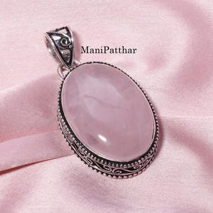 Puede incluir: Un colgante ovalado de cuarzo rosa engastado en un marco decorativo de plata de ley con intrincados detalles metálicos en los bordes y una anilla a juego. El texto ManiPatthar es visible cerca de la parte superior de la piedra preciosa rosa, lisa y pulida.