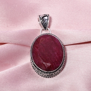 May include: A silver pendant with an oval-shaped ruby gemstone. The pendant has an intricate, ornate design around the gemstone.