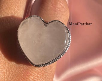 Pode incluir: Um anel com uma grande pedra rosa pálido em forma de coração, montada numa banda de metal prateado com um detalhe de rebordo com contas. O texto ManiPatthar é visível no lado direito do anel. A pedra tem uma superfície lisa e polida.