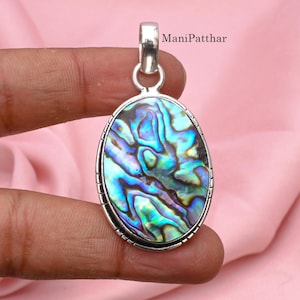 May include: An oval pendant featuring an iridescent abalone shell inlay set in a polished silver frame with a decorative bail. The surface displays swirling patterns of blue, green, and purple hues. The text ManiPatthar is visible in the corner.