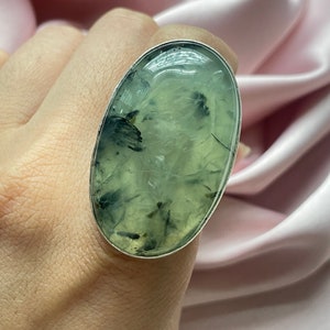 May include: A silver ring with a large oval green and black gemstone. The gemstone has a cloudy, translucent appearance with dark green and black inclusions.