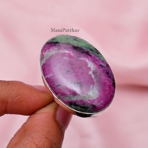 Op de afbeelding: Een ovaalvormige ring met een grote, gepolijste robijn-zoisiet edelsteen met opvallende magenta, witte en donkergroene minerale insluitingen. De steen is gezet in een gladde, gepolijste zilverkleurige metalen band. De tekst ManiPatthar is zichtbaar.
