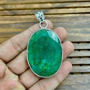 May include: An oval-shaped pendant featuring a large, faceted green emerald gemstone set in a silver frame with a decorative bail. The stone displays natural inclusions and a vibrant green hue, securely held by a beaded silver border.