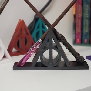 May include: A black triangular stand with a symbol of a triangle with a circle inside, holding three wands. The wands are made of wood and have different colored tips. The stand is on a white surface.