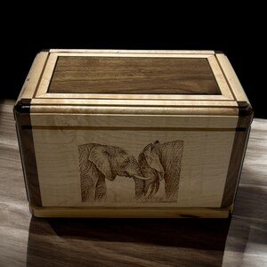 以下が含まれることがあります： クルミの天板とメープル製の側面を持つ木製の箱。箱の前面には、レーザー彫刻された2頭の象の画像が施されています。