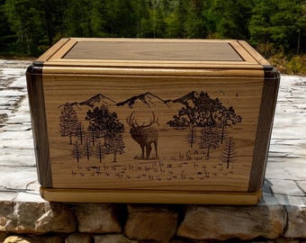 May include: A wooden box with a hinged lid, featuring a laser-engraved design of a mountain scene with an elk standing in the foreground. The box is made of light and dark wood, creating a contrasting effect.